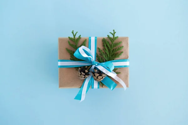 ギフト ボックスのペーパー クラフト、青と白のリボンと飾られたモミの枝と青い背景、上面に松ぼっくりをラップします。クリスマス プレゼント. — ストック写真