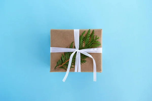 Подарункова коробка, обгорнута ремісничим папером, білою стрічкою та прикрашена ялиновим гілкою на синьому фоні, вид зверху. різдвяний подарунок . — стокове фото
