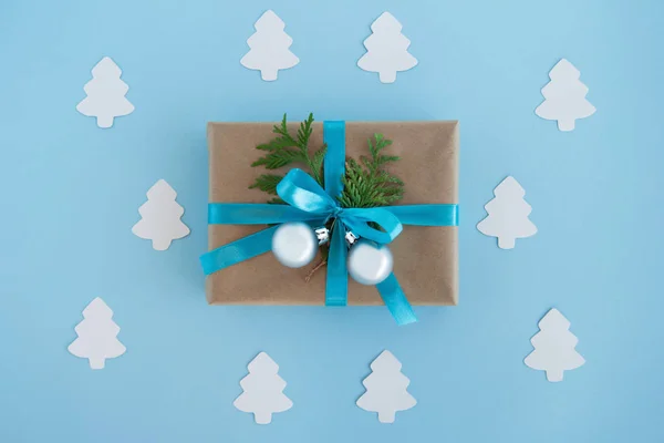 Luxe-geschenketui van Kraft papier, blauw lint gewikkeld en ingerichte fir takken en zilveren kerstballen met Witboek fir tree rond op de blauwe achtergrond, bovenaanzicht. — Stockfoto