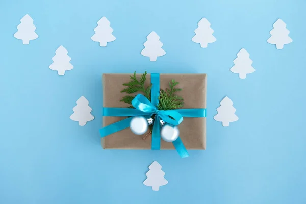 Luxe-geschenketui van Kraft papier, blauw lint gewikkeld en ingerichte fir takken en zilveren kerstballen op de blauwe achtergrond met Witboek fir tree, bovenaanzicht. — Stockfoto