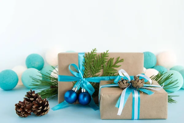 Caixas de presente de Natal embrulhadas de papel artesanal, fitas azuis e brancas, decoradas de ramos de abeto, cones de pinheiro e bolas de Natal no fundo azul e branco com luzes de Natal . — Fotografia de Stock