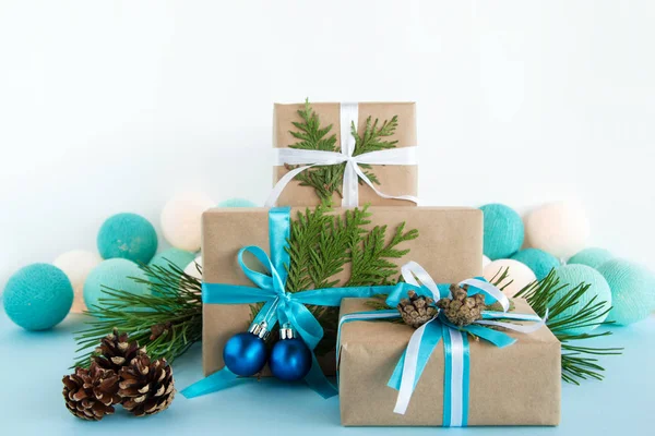 Drie kerst geschenkdozen verpakt van Kraft papier, blauwe en witte linten, ingericht van Spar takken, pine kegels en kerstballen op de blauwe en witte achtergrond met kerstverlichting. — Stockfoto