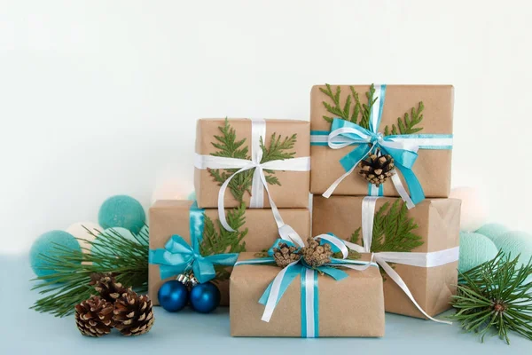 Vijf kerst geschenkdozen verpakt van Kraft papier, blauwe en witte linten, ingericht van Spar takken, pine kegels en kerstballen op de blauwe en witte achtergrond met kerstverlichting. — Stockfoto