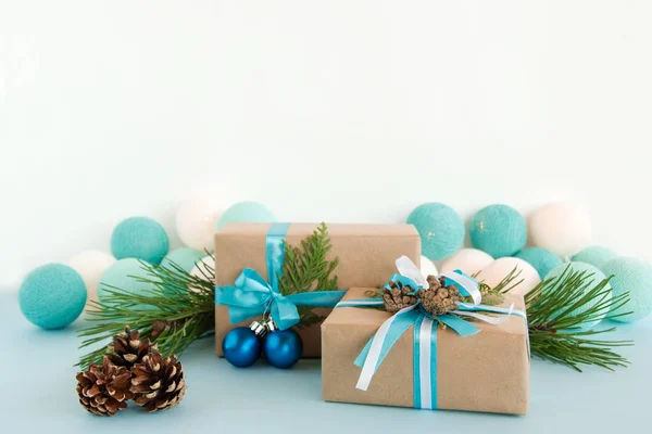 Twee kerst geschenkdozen verpakt van Kraft papier, blauwe en witte linten, ingericht van Spar takken, pine kegels en kerstballen op de blauwe en witte achtergrond met kerstverlichting. — Stockfoto