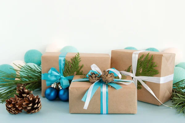 Drie kerst geschenkdozen verpakt van Kraft papier, blauwe en witte linten, ingericht van Spar takken, pine kegels en kerstballen op de blauwe en witte achtergrond met kerstverlichting. — Stockfoto