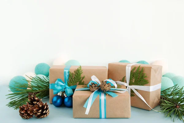 Drie kerst geschenkdozen verpakt van Kraft papier, blauwe en witte linten, ingericht van Spar takken, pine kegels en kerstballen op de blauwe en witte achtergrond met kerstverlichting. — Stockfoto