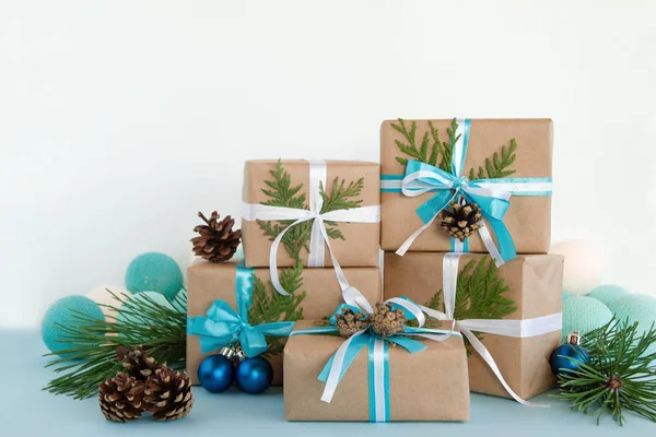 Різдвяні подарункові коробки, обгорнуті ремісничим папером, синьо-білими стрічками на синьо-білому тлі, прикрашені сосновими шишками, ялиновими гілками, різдвяним м'ячем та вогнями . — стокове фото
