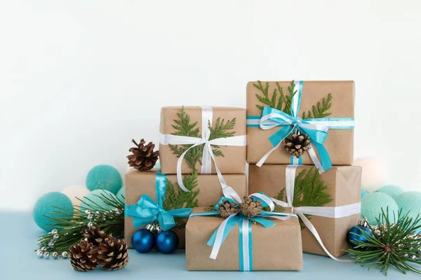 Caixas de presente de Natal embrulhadas de papel artesanal, fitas azuis e brancas sobre o fundo azul e branco decorado de cones de pinho, ramos de abeto, contas de prata, bola de Natal e luzes . — Fotografia de Stock