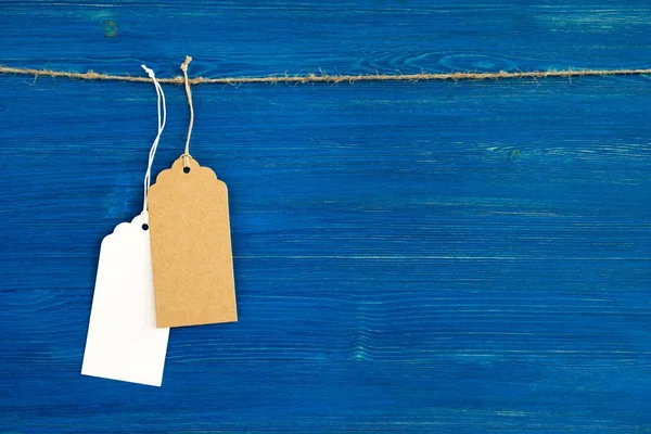 Dos etiquetas de precio de papel en blanco y marrón o etiquetas colgadas de una cuerda en el fondo azul de madera . — Foto de Stock