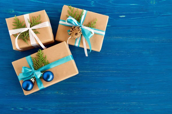 Подарункові коробки, обгорнуті ремісничим папером, синьо-білою стрічкою та прикрашені ялиновими гілками, сині різдвяні кульки та сосновий конус на синьому дерев'яному тлі, вид зверху. різдвяний подарунок . — стокове фото