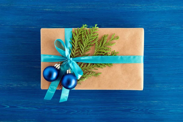 Luxe-geschenketui van Kraft papier, blauw lint gewikkeld en ingericht fir takken en blauwe kerstballen op de blauwe houten achtergrond, bovenaanzicht. Kerstcadeau. — Stockfoto