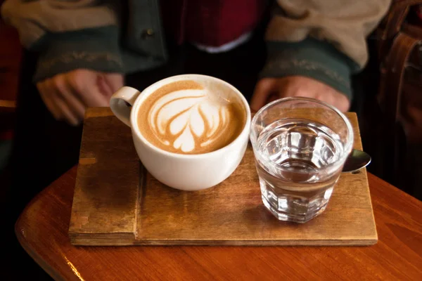 咖啡馆里的木桌上有一杯热卡布奇诺和水杯 — 图库照片