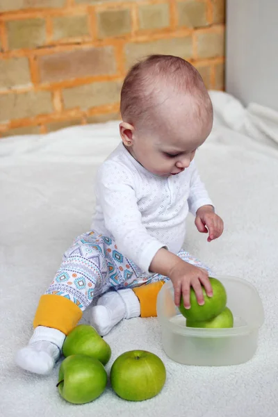 1歳の女の子は白いプレイでリビングルームに座っているとプラスチック製の容器に緑のリンゴで遊んでいます — ストック写真