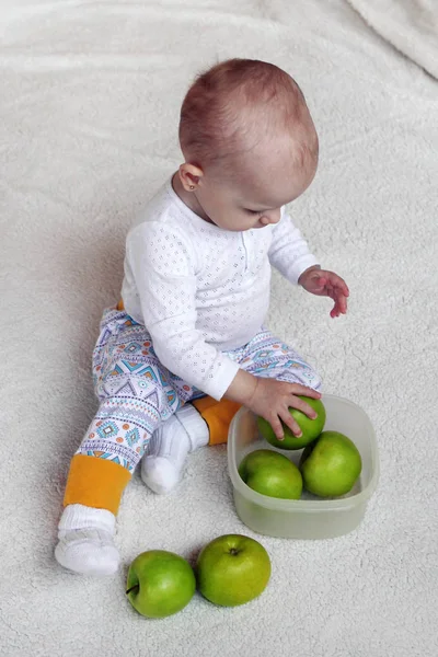 小さな女の子は白いプレイに座っているとプラスチック製の容器に緑のリンゴと遊ぶ — ストック写真