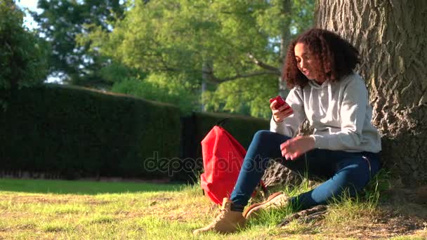 4 k-videoclip van mooie gezonde gemengd ras African American girl tiener zitten door een boom met een rode rugzak met behulp van een mobiele telefoon sms-tekstberichten — Stockvideo