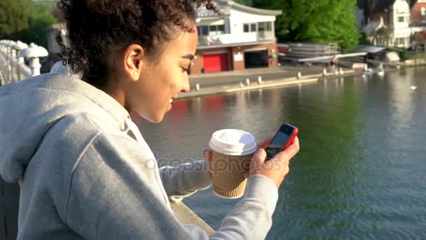 4K clipe de vídeo de bela raça mista afro-americana menina adolescente mulher em uma ponte sobre um rio, beber café takeout e usando um aplicativo de telefone celular ou mensagens de texto — Vídeo de Stock