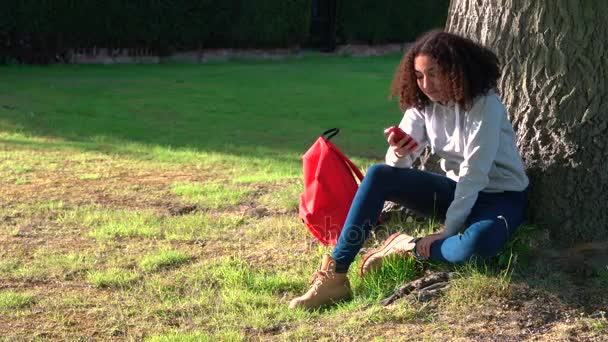 4 k-videoclip van mooie gezonde gemengd ras African American girl tiener zitten door een boom met een rode rugzak met behulp van een mobiele telefoon sms-tekstberichten — Stockvideo