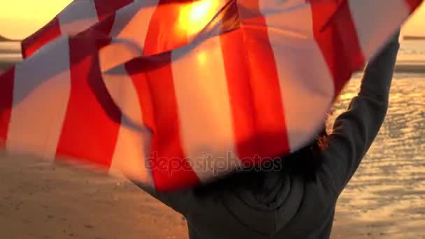 4K video clip de raza mixta chica afroamericana adolescente mujer joven sosteniendo una bandera estadounidense de estrellas y rayas en una playa al atardecer o al amanecer — Vídeo de stock