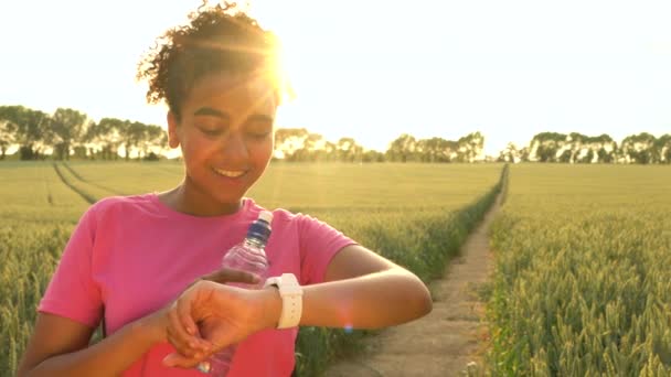 4k Videoclip von schönen fit gemischtes Rennen afrikanisch amerikanische Mädchen Teenager weibliche junge Läuferin mit Smart Watch und läuft auf Pfad durch Feld von Gerste oder Weizen bei Sonnenuntergang — Stockvideo