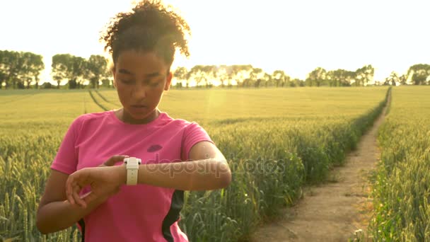 4 k-videoclip voor mooie fit gemengd ras African American girl tiener vrouwelijke jonge vrouw runner met behulp van slimme horloge en draait op pad door het veld van gerst of tarwe gewassen bij zonsondergang — Stockvideo