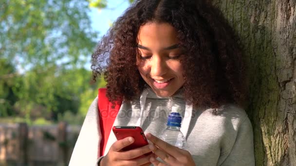 Güzel mutlu karışık yarış Afro-Amerikan Kız genç bir su şişesinden içiyor ve bir cep telefonu uygulaması veya metin ileti kullanarak kırmızı sırt çantasıyla karşı bir ağaca yaslanmış — Stok video