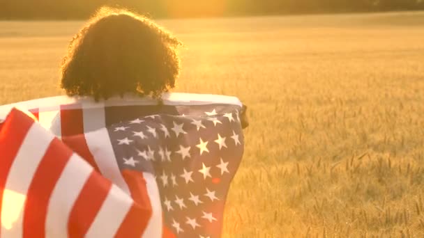 4K video clip de raza mixta chica afroamericana adolescente mujer joven sosteniendo una bandera estadounidense de estrellas y rayas en un campo de trigo al atardecer o al amanecer — Vídeo de stock