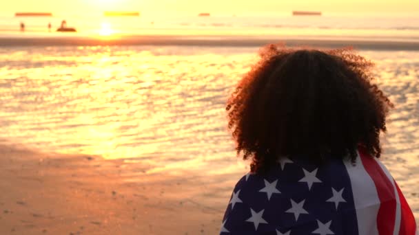 4K video clip de raza mixta chica afroamericana adolescente mujer joven envuelta en una bandera estadounidense de estrellas y rayas en una playa al atardecer o al amanecer — Vídeo de stock