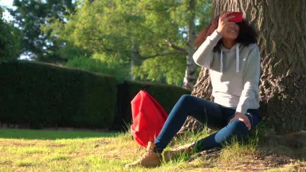 混血アフリカ系アメリカ人の女の子 10 代の赤いバックパックの木にもたれて、携帯電話のカメラを使用して — ストック動画
