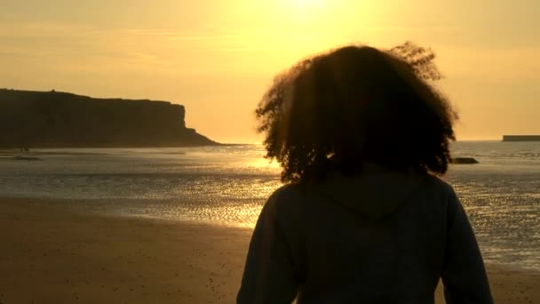 4K clipe de vídeo de triste pensativo misto raça afro-americana menina adolescente mulher jovem com cabelo encaracolado em pé em uma praia olhando para o pôr do sol ou nascer do sol — Vídeo de Stock