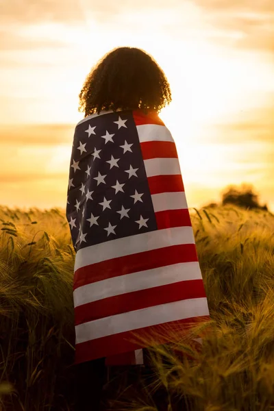 Meisje tiener vrouw gewikkeld in Usa vlag in veld bij zonsondergang — Stockfoto