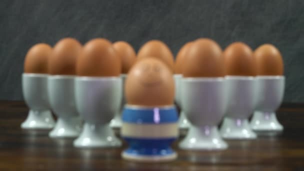 木のテーブルの上の白い卵のカップに卵のグループの前で笑顔でストライプの卵カップでゆで卵のラックフォーカスショット 異なるされています 個性の概念 — ストック動画