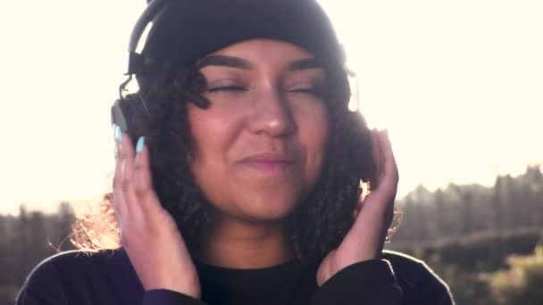 美丽的混血非洲裔美国少女 头戴黑色贝尼帽 在夕阳西下 在太阳光的映照下 在耳机里听着流淌的音乐 — 图库视频影像