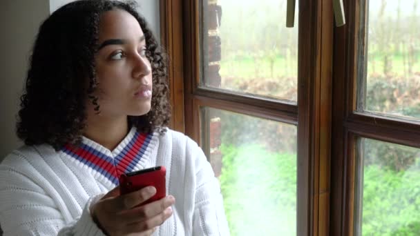 悲しい美しい混合レースアフリカ系アメリカ人の女の子ティーン若いです女性は白いプルオーバーを身に着けています 彼女の携帯電話やスマートフォンを使用してウィンドウによって座っているソーシャルメディア 通信やテキストメッセージのために — ストック動画