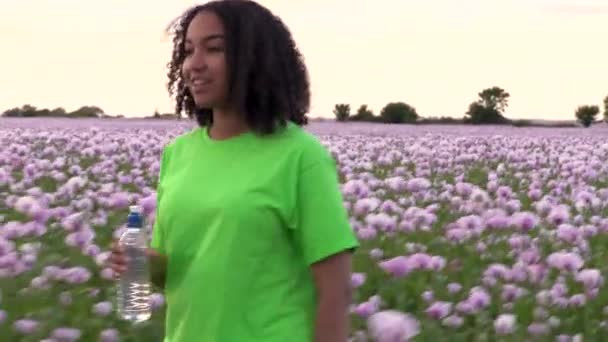 美しい幸せな混合レースアフリカ系アメリカ人の女の子ティーン女性若いです女性のフィールドを歩くピンクケシの花飲料水からプラスチックボトル — ストック動画