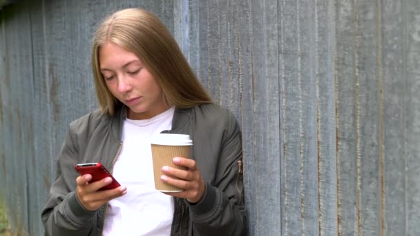 漂亮的金发少女用她的智能手机或手机社交媒体 喝着外卖咖啡 — 图库视频影像