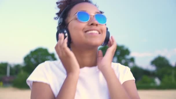 スローモーションフォロービデオの混合レースアフリカ系アメリカ人の女の子ティーン若いです女性身に着けていますAホワイト シャツと青サングラス歩くと音楽を聞いて彼女の携帯電話とワイヤレスヘッドフォン — ストック動画