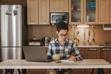 Bir kadının çalıştığı yemek masası. Uzaktan kumandalı sitede ev ödevi. Rahat kıyafetler, evin rahatlığı.