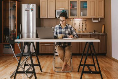 Bir kadının çalıştığı yemek masası. Uzaktan kumandalı sitede ev ödevi. Rahat kıyafetler, evin rahatlığı.