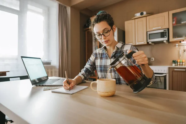 A woman pours tea into a Cup and works at remote home work. Quarantine, self-isolation