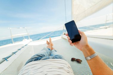 Bir yattaki bir tatilcinin elinde telefon var. Tatilin boyunca temasta kal. Hızlı internet. Telefonundaki içeriğe dikkat et. Diğer ülkelerde dolaşmak.