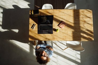 Evde çalışan ve kamerada gülümseyen bir kadın. Üst Manzara. Parlak ışık ve uzun gölgeli bir oda