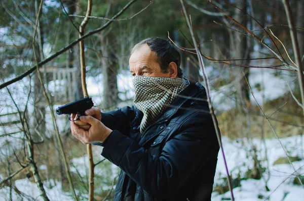 An elderly man with a gun in the woods in early spring. Man dressed in a black jacket. The face of unknown covered by a scarf. He holds the gun in both hands and aiming into the distance