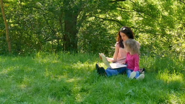 Щасливі маленька дівчина Prepears увійти до початкової школи з її тітка, сидячи на свіжі зелена трава у міському парку і використовуючи білий ноутбук. — стокове відео