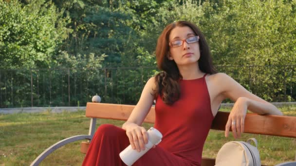 Retrato al aire libre de la atractiva morena vestida con anteojos sentada en el banco del parque y bebiendo algo de bebida de la Thermo Cup . — Vídeos de Stock