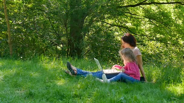 La niña inteligente se prepara para entrar en el jardín de infantes con su tutor. Mujer joven explica cómo usar el ordenador portátil para una niña rubia sentada en la hierba verde del parque . — Vídeos de Stock
