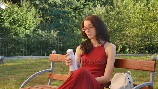 Linda adolescente con vestido largo rojo oscuro y anteojos bebiendo algo de la taza termo sentada en el banco al aire libre y mostrando el dedo grande . — Vídeos de Stock