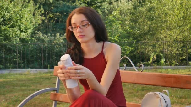 Belle fille aux cheveux foncés dans les lunettes de vue assis sur le banc en plein air avec bouteille de boisson et boire quelque chose pendant la journée chaude d'été . — Video