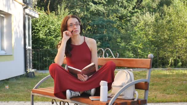 Junge Brünette mit Brille, die draußen auf dem Brunnen sitzt und etwas schreibt. Studentin im roten Kleid lernt draußen an sonnigen, warmen Tagen im Park. — Stockvideo