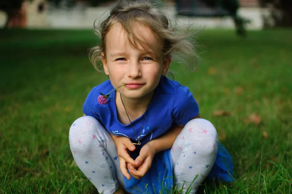 Bebek çömelme ve kameraya bakıyor. Kızlar gözleri ironik daralmış. O ağzına bir mor yonca çiçek tutan.