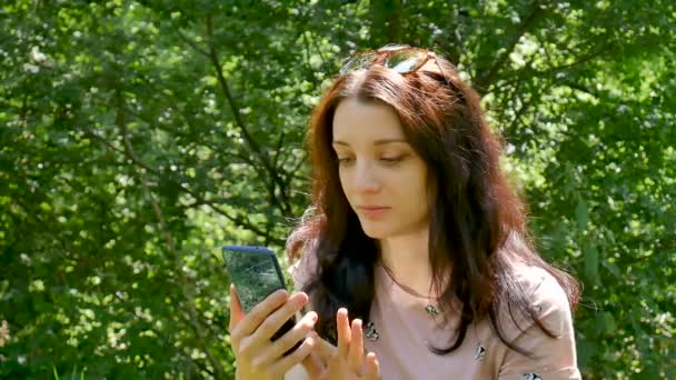 Erstaunliches Mädchen lacht, wenn sie Nachrichten in sozialen Netzwerken auf ihrem Handy liest. Nahaufnahme Porträt der schönen brünetten Frau mit blauem Smartphone im Freien auf grünen Bäumen Hintergrund. — Stockvideo